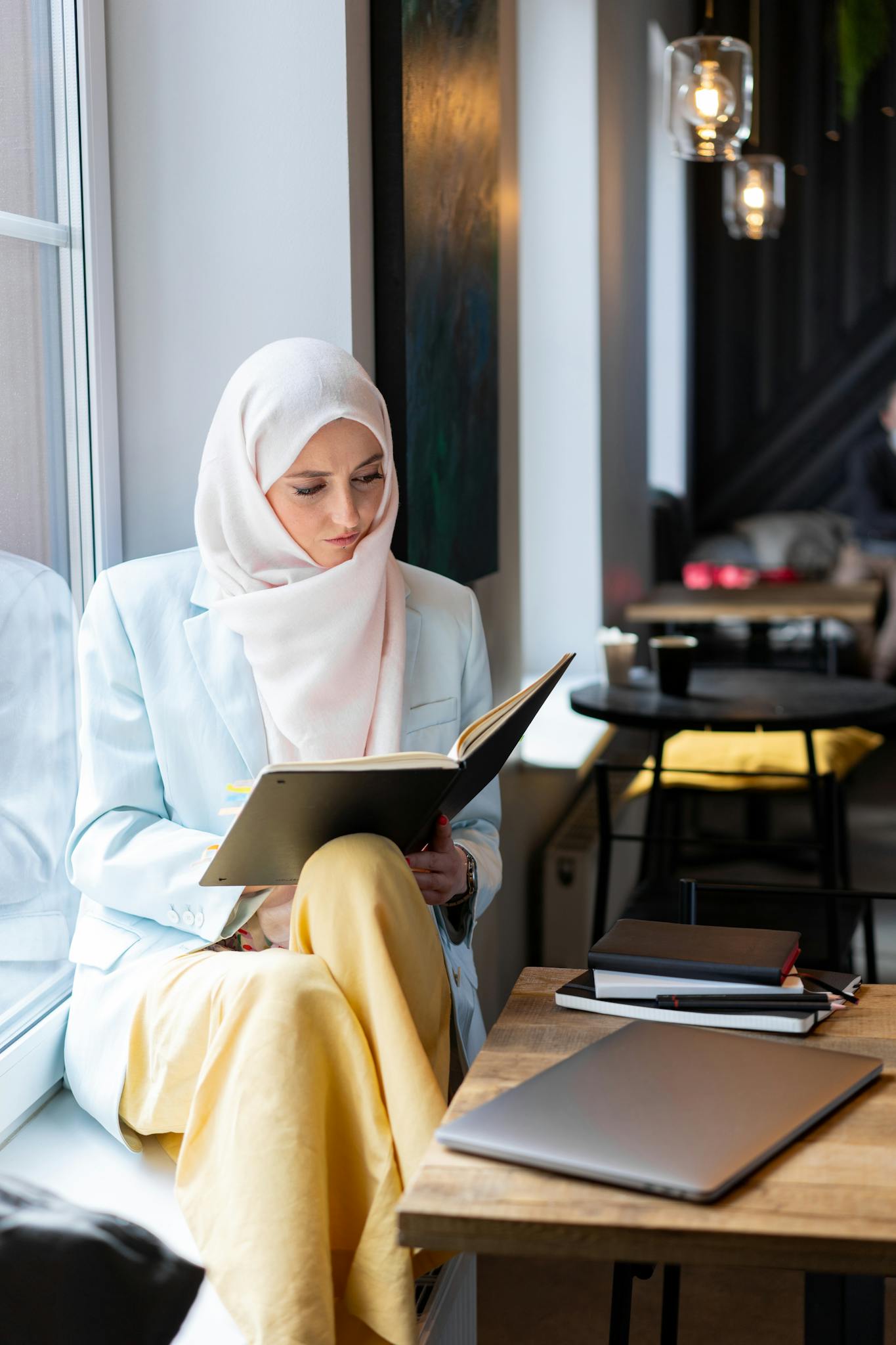 A young Muslim woman in a hijab reading a book at a modern cafe, with a laptop on the table.